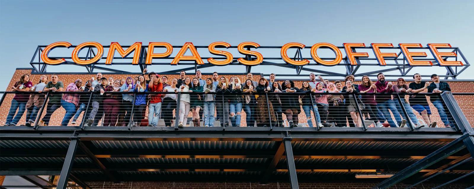 The Compass Coffee team waving from the roastery balcony under the Compass Coffee sign